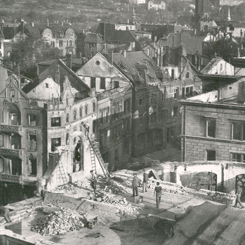 Auf einem Schwarzweißfoto sind mehrere Menschen zu sehen, die inmitten von Trümmern und beschädigten Gebäuden in einer Stadt stehen. Teilweise zerstörte Dächer und Fassaden sind sichtbar, was auf schwere Zerstörungen hindeutet, die vermutlich auf den Krieg zurückzuführen sind.