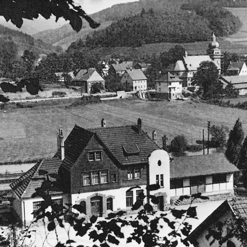 Schwarzweißfoto eines kleinen europäischen Dorfes mit Häusern, offenen Feldern und einer Kirche mit Kirchturm, umgeben von bewaldeten Hügeln und teilweise eingerahmt von Ästen im Vordergrund.