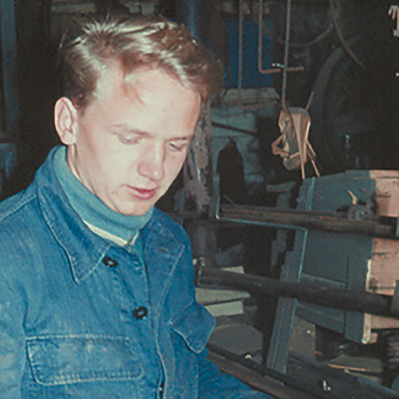 Ein junger Mann mit hellem Haar und einer blauen Jeansjacke blickt nach unten, während er in einer Industrie- oder Werkstattumgebung mit Maschinen und Werkzeugen im Hintergrund steht.