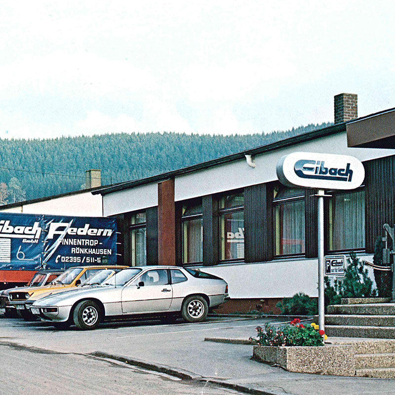 Ein silberner Oldtimer-Sportwagen parkt vor einem niedrigen, modernen Industriegebäude mit Eibach-Schildern. Im Hintergrund sind unter einem bewölkten Himmel kiefernbedeckte Hügel zu sehen.