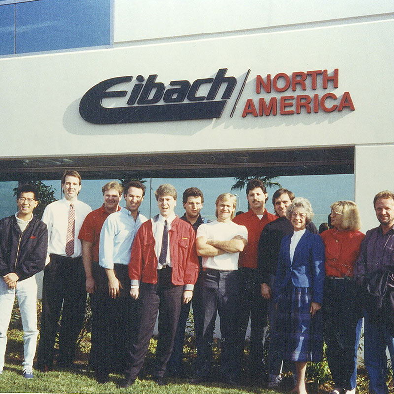 Eine Gruppe von zwölf Personen steht lächelnd vor einem Gebäude mit einem großen Eibach North America-Schild. Die Gruppe besteht aus Männern und Frauen in Business- und Freizeitkleidung.