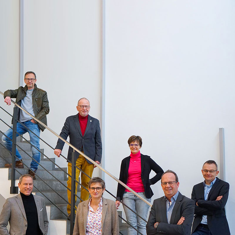 Sieben Erwachsene posieren im Innenbereich; vier stehen am Fuß einer Treppe, drei stehen auf der Treppe. Sie tragen Business-Casual-Kleidung und lächeln vor einer schlichten weißen Wand.