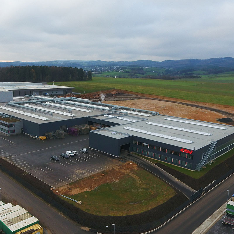 Luftaufnahme eines großen, modernen Industriegebäudes mit grauem Dach, umgeben von Parkplätzen, einigen geparkten Autos und offenen Grasfeldern, mit Hügeln und einer entfernten Stadt im Hintergrund.