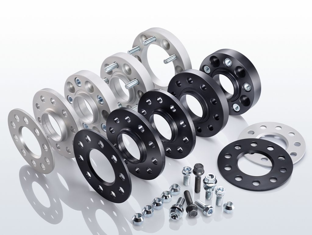 Ein Satz metallischer Spurverbreiterungen in Silber und Schwarz, in Reihen angeordnet, mit Schrauben und Muttern davor auf einer reflektierenden weißen Oberfläche.