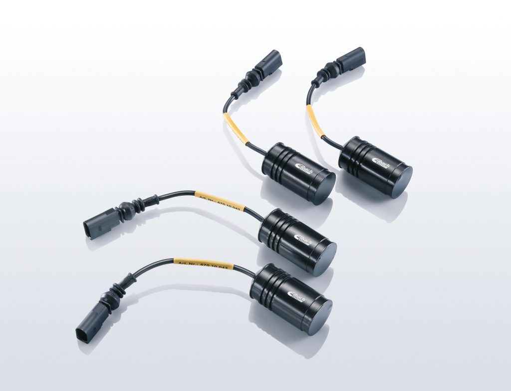 Vier schwarze zylindrische elektronische Komponenten mit angeschlossenen Anschlüssen und kurzen gelb beschrifteten Kabeln sind auf einer reflektierenden weißen Oberfläche angeordnet.