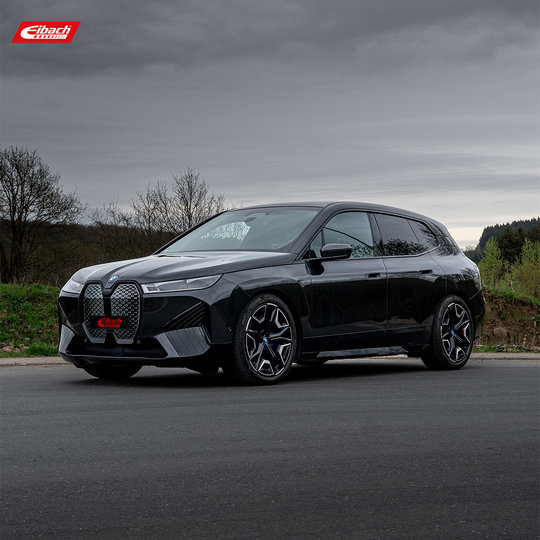 Ein schwarzer BMW SUV parkt auf einer asphaltierten Straße bei bedecktem Himmel. Das Auto zeichnet sich durch einen markanten Kühlergrill und sportliche Felgen aus. Im Hintergrund sind Bäume und Gras zu sehen. Das Eibach-Logo ist in der oberen linken Ecke zu sehen.