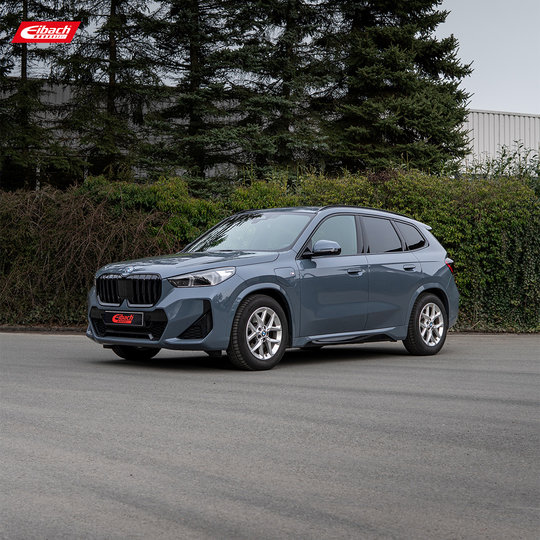 Vor grünen Büschen und hohen Bäumen parkt ein blauer BMW SUV auf einer befestigten Fläche. Im Hintergrund ist ein weißes Gebäude angeschnitten. Links oben ist das Eibach-Logo zu sehen. Vor grünen Büschen und hohen Bäumen parkt ein blauer BMW SUV auf einer befestigten Fläche. Im Hintergrund ist ein weißes Gebäude angeschnitten. Links oben ist das Eibach-Logo zu sehen.