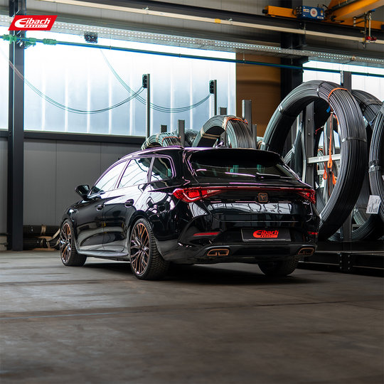 Ein eleganter schwarzer Kombi mit getönten Scheiben steht in der Nähe großer Industriekabeltrommeln. Das Auto hat polierte Felgen und das Eibach-Logo auf dem Nummernschild und in der oberen linken Ecke. Ein eleganter schwarzer Kombi mit getönten Scheiben steht in der Nähe großer Industriekabeltrommeln. Das Auto hat polierte Felgen und das Eibach-Logo auf dem Nummernschild und in der oberen linken Ecke.