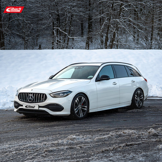 A white Mercedes-Benz station wagon with black alloy wheels is parked on a wet road in front of a snow-covered landscape and trees. The Eibach logo is visible in the top left corner. A white Mercedes-Benz station wagon with black alloy wheels is parked on a wet road in front of a snow-covered landscape and trees. The Eibach logo is visible in the top left corner.