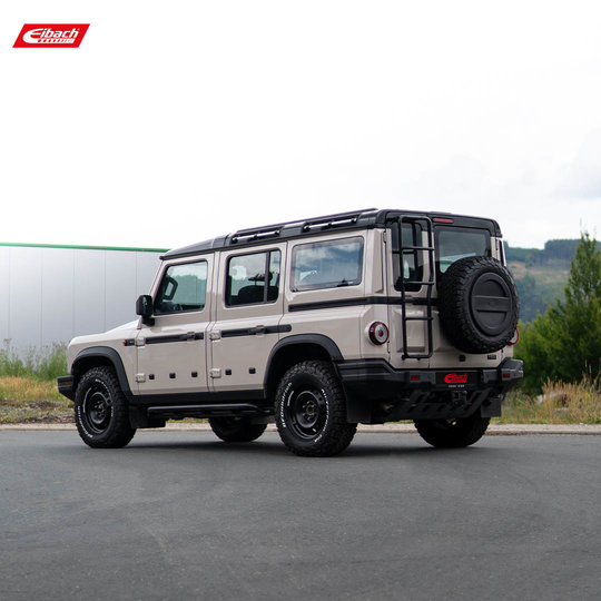 A beige off-road SUV with a roof rack and a rear-mounted spare tire is parked on a paved surface, with greenery and a building in the background. The Eibach logo is visible in the top left corner. A beige off-road SUV with a roof rack and a rear-mounted spare tire is parked on a paved surface, with greenery and a building in the background. The Eibach logo is visible in the top left corner.