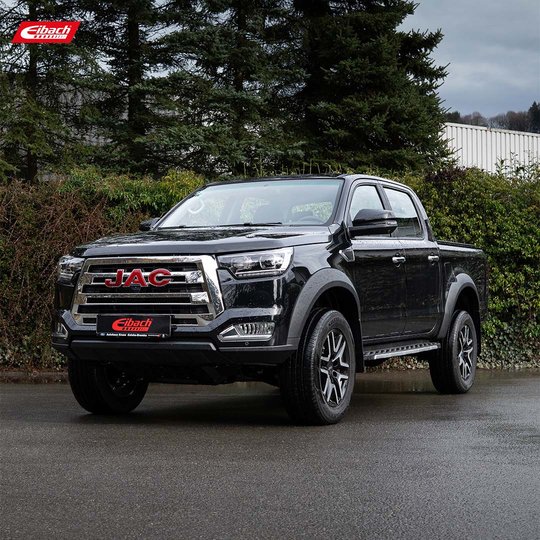 Ein schwarzer JAC Pickup parkt auf einer nassen Straße in der Nähe von grünen Büschen und Bäumen. Im Hintergrund ist ein Gebäude zu sehen. In der oberen linken Ecke ist das Eibach-Logo zu sehen. Ein schwarzer JAC Pickup parkt auf einer nassen Straße in der Nähe von grünen Büschen und Bäumen. Im Hintergrund ist ein Gebäude zu sehen. In der oberen linken Ecke ist das Eibach-Logo zu sehen.
