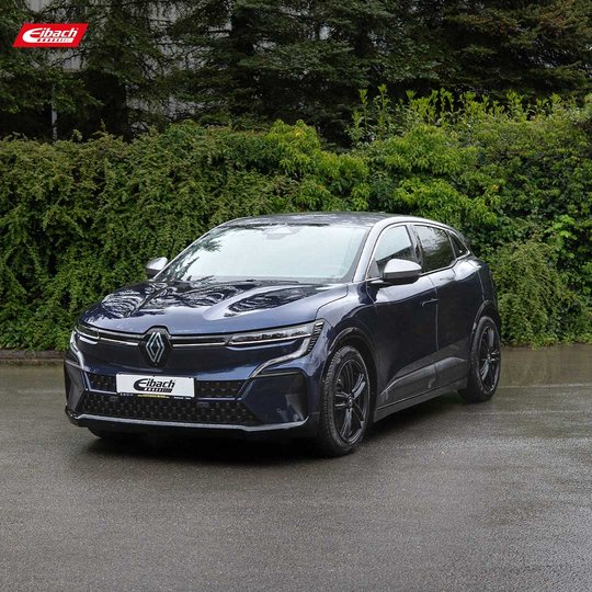 Ein dunkelblaues Elektrofahrzeug von Renault parkt auf einer nassen Straße, umgeben von üppigen grünen Büschen und Bäumen im Hintergrund. Das Auto zeichnet sich durch ein schlankes, modernes Design und schwarze Räder aus.