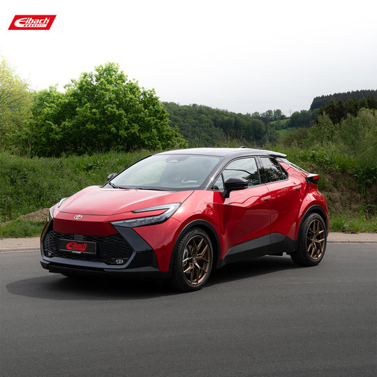 A shiny red compact crossover car with bronze alloy wheels is parked on a paved road, surrounded by green trees and hills under a cloudy sky. The Eibach logo is displayed in the top-left corner. A shiny red compact crossover car with bronze alloy wheels is parked on a paved road, surrounded by green trees and hills under a cloudy sky. The Eibach logo is displayed in the top-left corner.