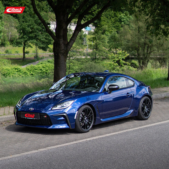 A blue sports car with black rims is parked on the side of a road, surrounded by greenery and trees, with a red Eibach logo in the top left corner of the image. A blue sports car with black rims is parked on the side of a road, surrounded by greenery and trees, with a red Eibach logo in the top left corner of the image.