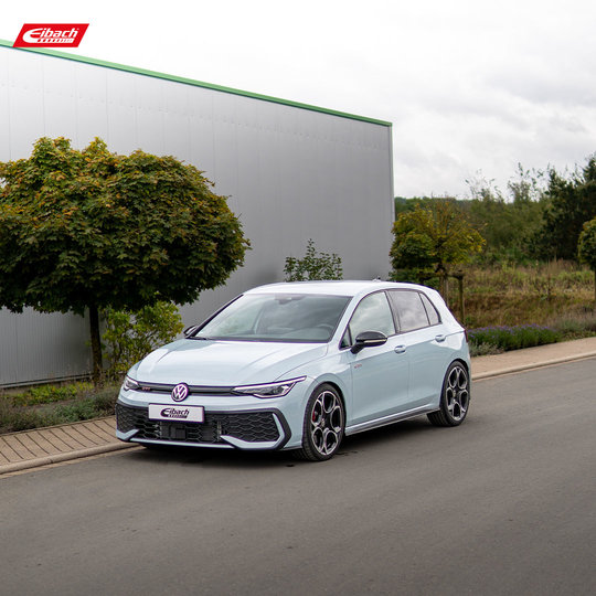 Ein weißer Volkswagen Golf Fließheck steht in einer ruhigen Straße neben Bäumen und einem großen Industriegebäude und hat in der oberen linken Ecke ein Eibach-Logo.