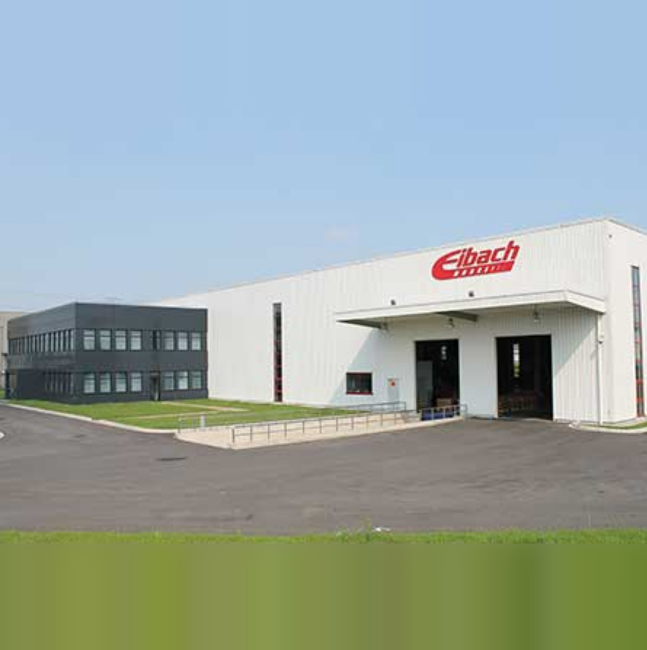 Ein großes weißes Industriegebäude mit dem roten Eibach-Logo über einem breiten Garagentor, daneben ein kleineres Bürogebäude, umgeben von gepflasterten Flächen und Gras unter einem klaren Himmel.