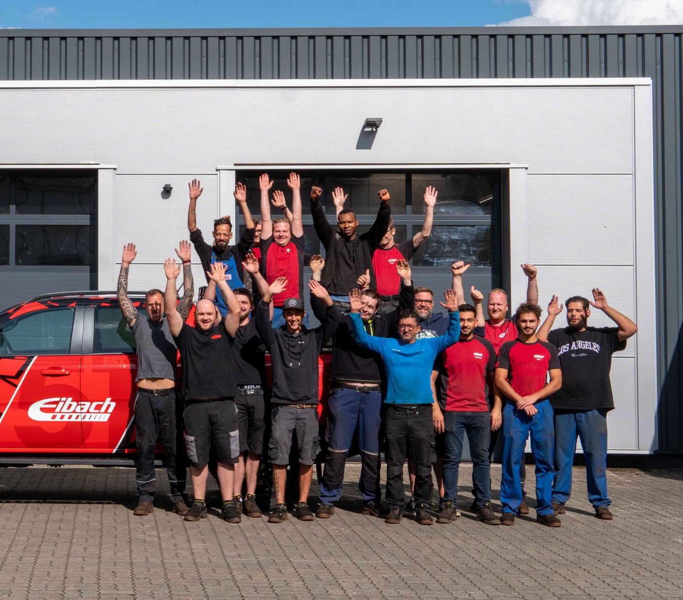 Eine Gruppe von sechzehn Personen steht im Freien vor einer Werkstatt. Einige tragen rote Hemden, alle lächeln und heben die Arme. Links parkt ein rotes Fahrzeug mit Eibach-Branding.