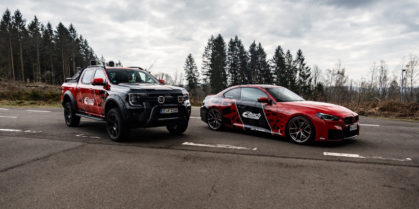 Ein schwarzer Pickup und ein roter Sportwagen mit Elbach-Branding parken auf einer leeren Straße, umgeben von Bäumen unter einem bewölkten Himmel.