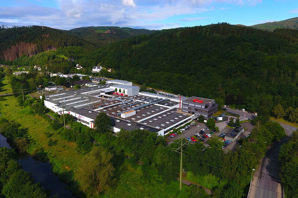 Eine große Industrieanlage mit weißen Dächern und einem roten Logo ist von dichtem grünen Wald und Hügeln umgeben. In der Nähe sind unter einem teilweise bewölkten Himmel ein Parkplatz mit Autos und eine Straße zu sehen.