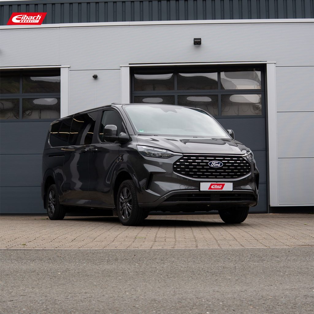 Ein schwarzer Ford Tourneo Custom ist vor einem großen grauen Industrietor geparkt. In der linken oberen Ecke ist das Eibach-Logo zu sehen, das auf die Höherlegung des Fahrzeugs nach einem erfolgreichen Probeaufbau hinweist.