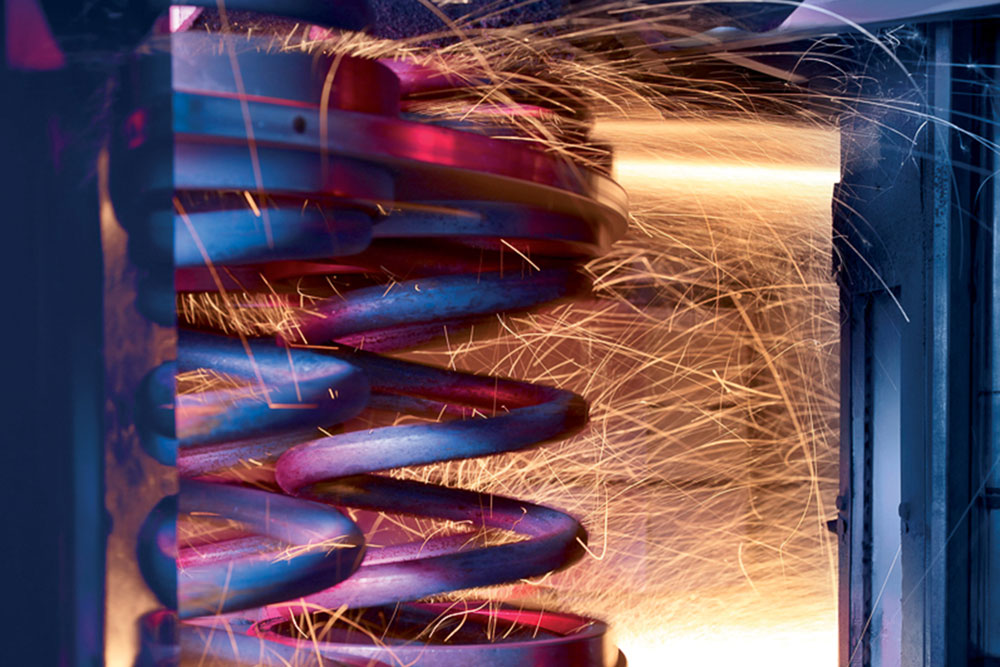 Nahaufnahme einer gewundenen Metallfeder mit leuchtend orangefarbenen Funken, die im Hintergrund fliegen und auf eine industrielle Metallbearbeitung oder ein laufendes Schleifverfahren hindeuten. Die Szene ist mit blauen und violetten Tönen beleuchtet und fängt einen dynamischen Moment aus der Perspektive der Leistungsübersicht ein.