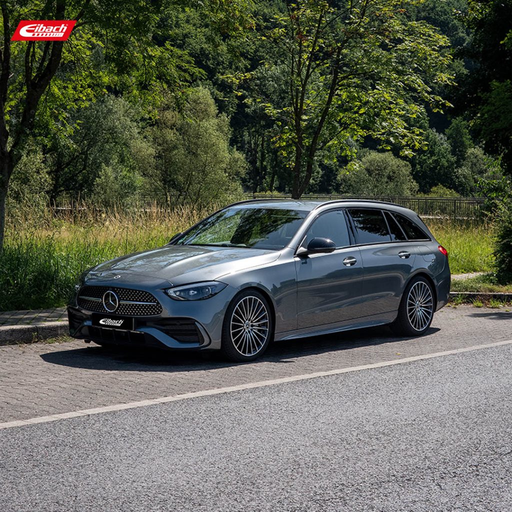 Ein schnittiger grauer Mercedes-Benz Kombi steht auf einem gepflasterten Straßenrand neben grünem Gras und Bäumen, unter einem klaren Himmel, mit dem Eibach-Logo in der oberen linken Ecke.