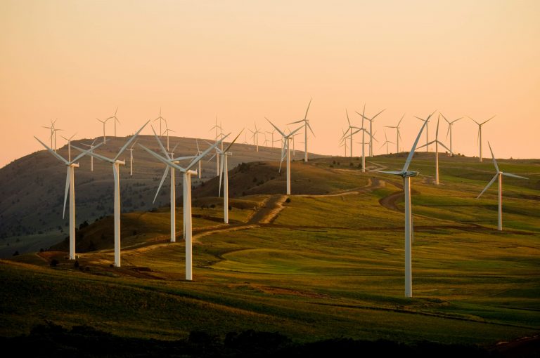 Windturbinen, verstreut über sanfte grüne Hügel bei Sonnenuntergang, sanftes orangefarbenes Licht am Himmel und verschlungene Feldwege, die sich durch die grasbewachsene Landschaft schlängeln - das sind die Highlights moderner Industrieinnovationen wie die Lösungen von Eibach Anwendungen Industrie.