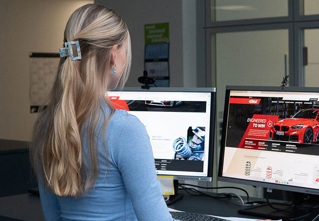 Eine Frau mit langen blonden Haaren in einem blauen Top arbeitet an einem Schreibtisch vor zwei Computermonitoren, auf denen Websites über Autos und Rennsport angezeigt werden, auf denen Eibach als Hersteller von maßgeschneiderten Federungslösungen vorgestellt wird, und das alles in einer professionellen Büroumgebung.