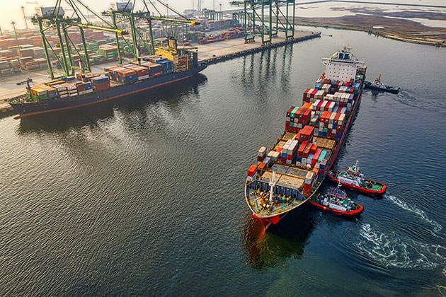 Ein großes, mit bunten Containern beladenes Frachtschiff wird von Schleppern durch einen belebten Hafen gelenkt und zeigt anwendungsbeispiele Industriefedern in Kränen und Hafenanlagen für einen effizienten Frachtumschlag.