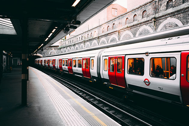 Ein Zug der Londoner U-Bahn mit roten und weißen Waggons hält an einem Außenbahnsteig. Die Fahrgäste sind im Inneren zu sehen, während die Backsteinmauer entlang der Gleise auf das industrielle Erbe Londons hinweist.