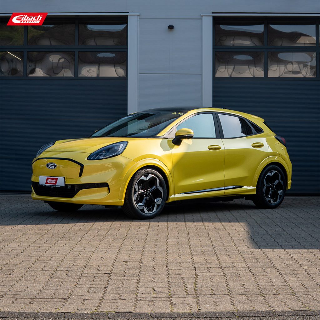 Ein leuchtend gelber Ford Puma parkt auf einer gepflasterten Fläche vor zwei großen geschlossenen Garagentoren, mit Eibach-Schriftzug in der linken oberen Ecke. In der Nähe sorgt ein Mercedes-Benz C 300 de (Baureihe 206) für einen Hauch moderner Eleganz in der sportlichen Szene.