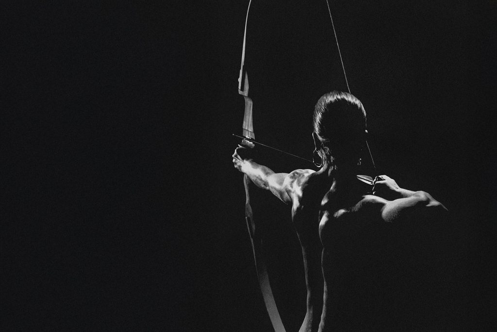 Eine muskulöse Person mit langem Haar, die von hinten in dramatischem Licht gesehen wird, spannt einen Bogen und zielt mit einem Pfeil in die Dunkelheit, was ihre Stärke und Konzentration unterstreicht.