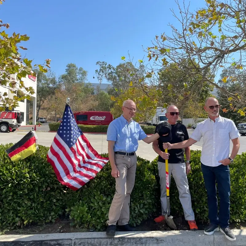 Drei Männer stehen im Freien in der Nähe eines Bordsteins, einer mit einer Schaufel in der Hand, und markieren Meilensteine der Eibach Firmengeschichte. Hinter ihnen sind deutsche und amerikanische Flaggen zu sehen, im sonnigen Hintergrund sind Bäume und Gebäude zu erkennen.