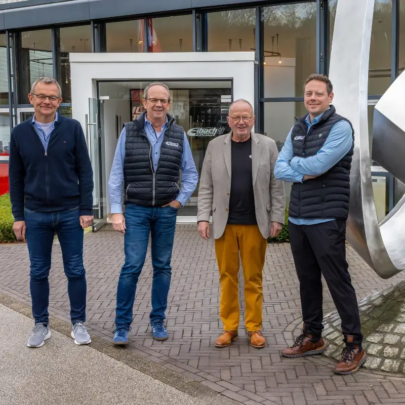 Vier Männer stehen vor einem modernen Gebäude mit großen Fenstern und lächeln neben einer großen Metallskulptur. Ihre legere Kleidung zeugt von Kameradschaft, während sie auf diesem Foto ein Stück eibach Firmengeschichte feiern.