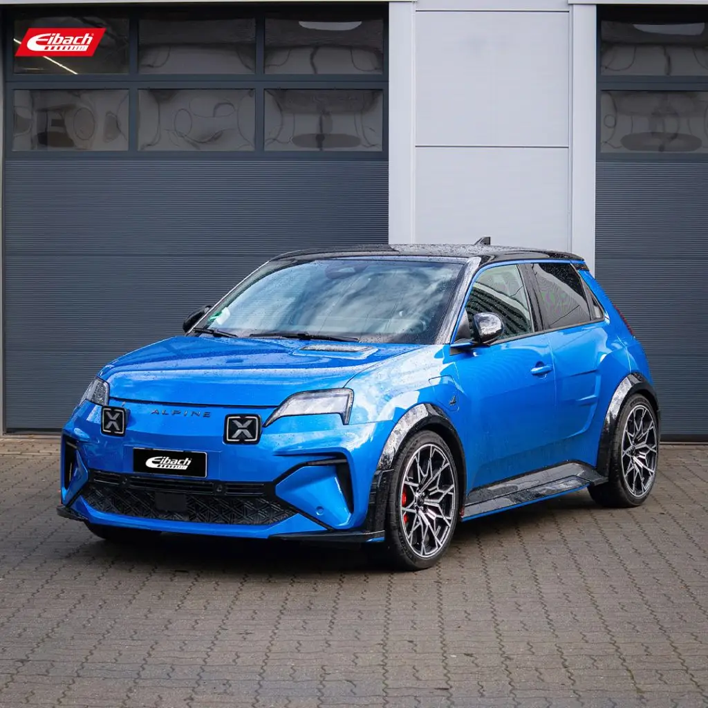 A bright blue Alpine A290 electric hatchback, equipped with a sport-tuned suspension setup and black lightweight alloy wheels, is parked on brick paving in front of a modern grey garage building. The performance-oriented stance and sharpened driving dynamics reflect its tuning focus. The Eibach logo is visible in the upper left corner.