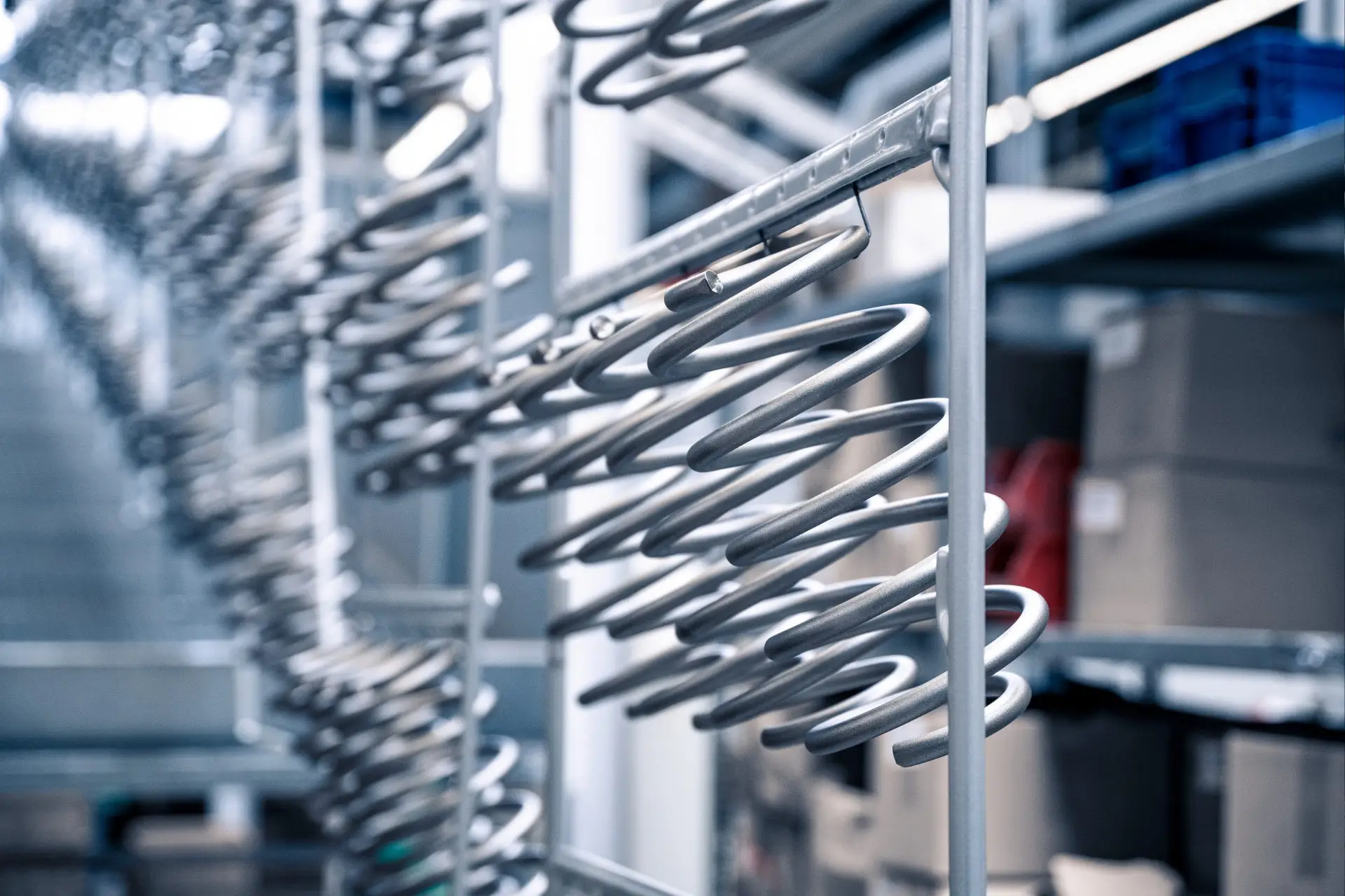 Nahaufnahme von großen Metallfedern oder -spulen, die in einer Reihe auf einem Regal in einem Industrielager angeordnet sind, wobei Regale und Kisten im Hintergrund verschwimmen.