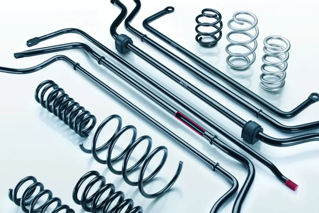 Eine Sammlung von Autoaufhängungsteilen aus Metall, darunter mehrere schwarze Stabilisatoren, schwarze Spiralfedern und einige silberne Spiralfedern, alle ordentlich auf einer weißen Fläche angeordnet.