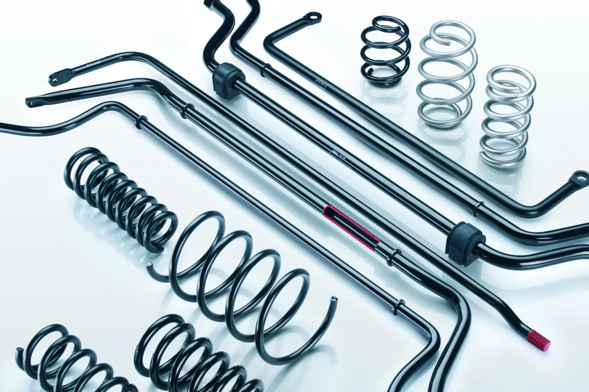 Eine Sammlung von Autoaufhängungsteilen aus Metall, darunter mehrere schwarze Stabilisatoren, schwarze Spiralfedern und einige silberne Spiralfedern, alle ordentlich auf einer weißen Fläche angeordnet.