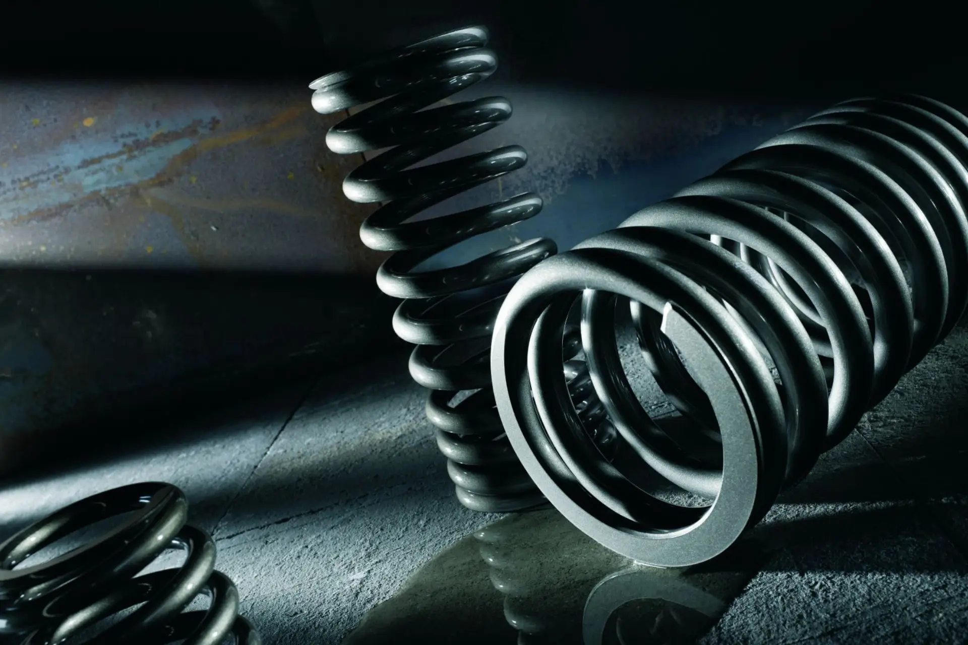 Nahaufnahme von drei großen, metallischen Spiralfedern auf einer strukturierten, grauen Oberfläche mit dramatischen Licht- und Schatteneffekten. Der Hintergrund ist dunkel und industriell und hebt die glänzenden, glatten Oberflächen der Federn hervor.