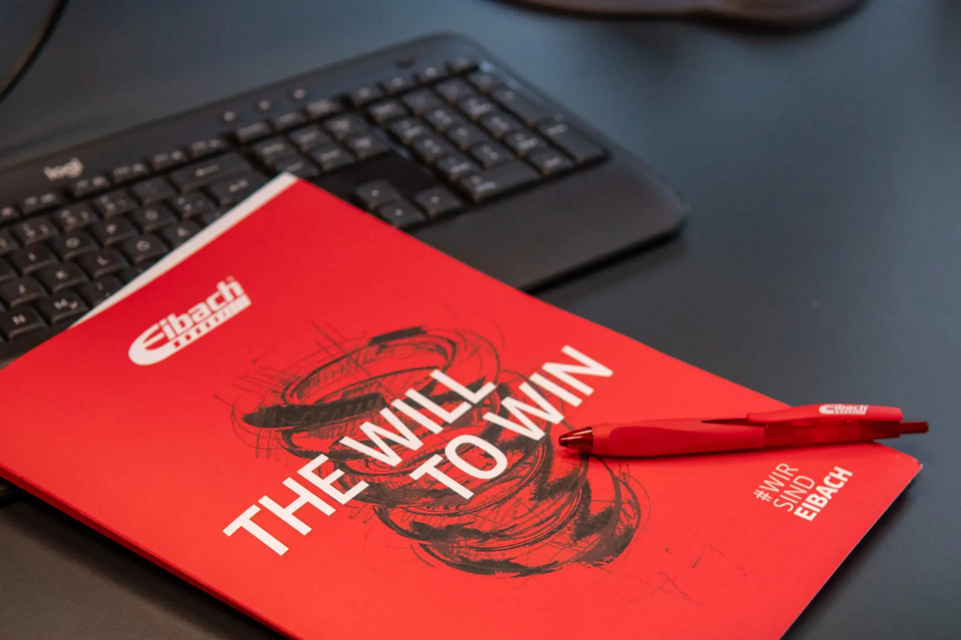 Ein rotes Büchlein mit dem Titel THE WILL TO WIN und dem Eibach-Logo liegt auf dem Schreibtisch, daneben ein roter Stift und eine schwarze Computertastatur.