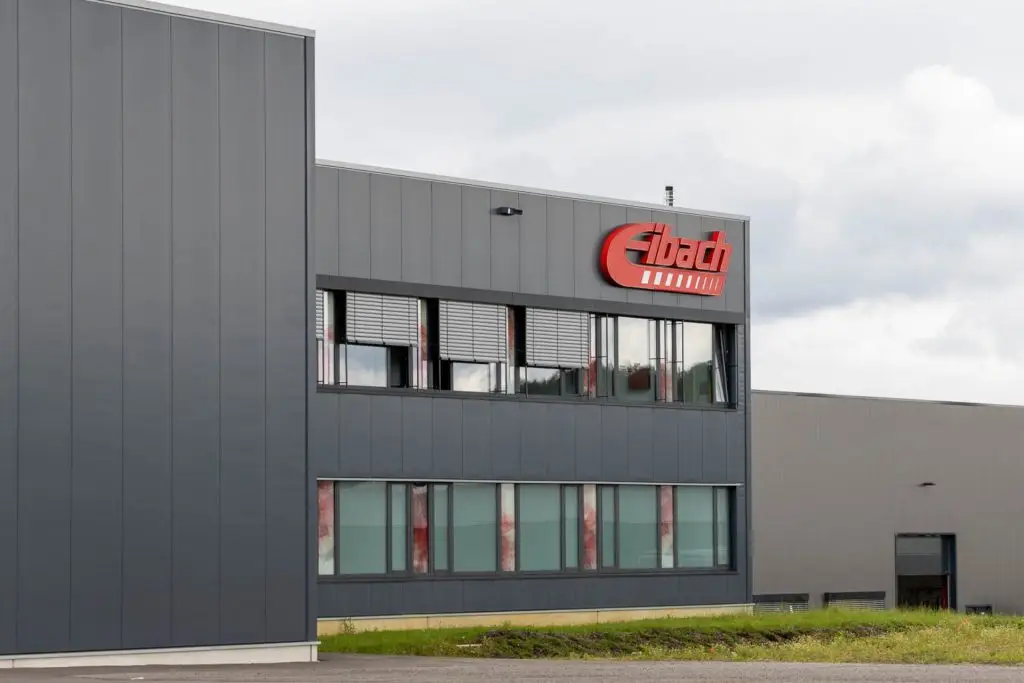 Modernes Industriegebäude mit dem Eibach-Logo in Rot auf der oberen Fassade, mit großen Fenstern und grauen Außenverkleidungen vor einem bewölkten Himmel.
