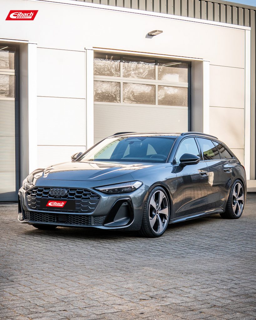 Ein grauer Audi A5 Avant Kombi mit sportlichen Felgen, ausgestattet mit Sportline-Tieferlegungsfedern, steht auf einem gepflasterten Parkplatz vor einem modernen Gebäude mit großen Garagentoren und Eibach-Logos – ein beeindruckendes Anwendungsbeispiel für eine Fahrwerkskonfiguration.