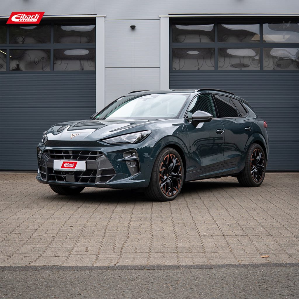 Ein dunkelgrüner Cupra-SUV steht auf einer gepflasterten Fläche vor modernen, grauen Garagentoren. Das Auto hat sportliche schwarze Felgen und ein Eibach-Kennzeichen. Das Eibach-Logo ist in der oberen linken Ecke zu sehen.